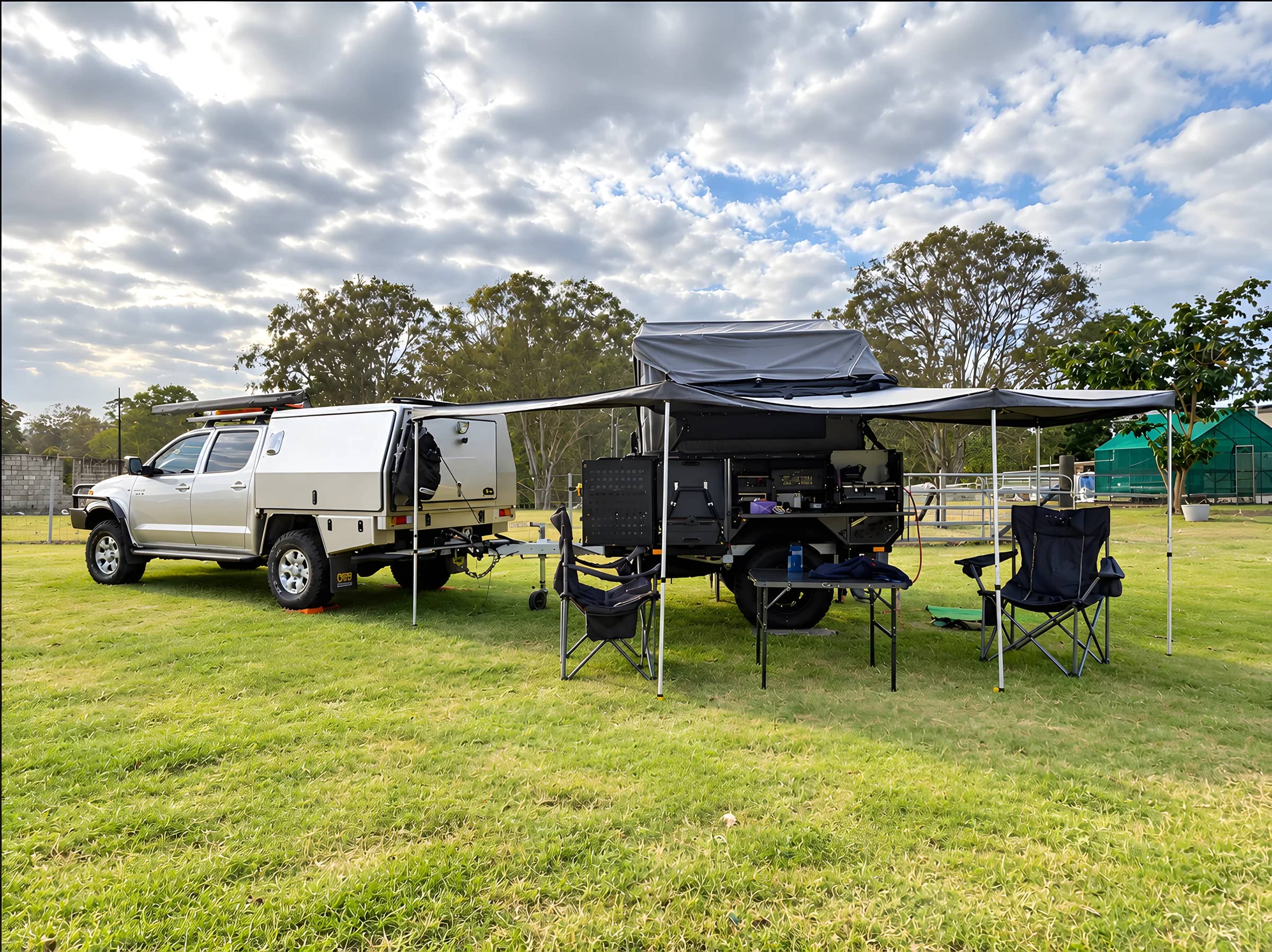 A ute and camper trailer set up under an awning on a grassy campsite, with camp chairs and a table